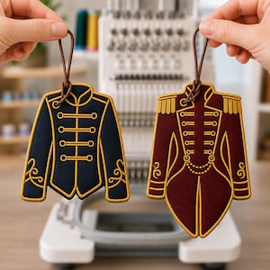 May include: Two embroidered ornaments shaped like military-style jackets. One is dark navy blue with gold trim and horizontal toggles. The other is deep burgundy with gold epaulets, toggles, and a decorative chain detail. Both have brown hanging loops.