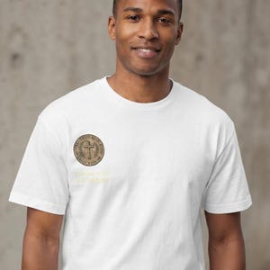 Puede incluir: Una camiseta blanca de manga corta y cuello redondo con un emblema circular marrón en el pecho izquierdo. El emblema muestra una cruz rodeada de hojas de laurel y texto. Debajo, el texto amarillo dice FINDING YOUR BEST VERSION.