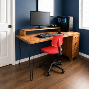 May include: A wooden computer desk with a monitor stand, featuring a natural wood grain finish, black metal hairpin legs, and a three-drawer storage unit on the right side. A bright red office chair with a perforated backrest sits in front of the desk.