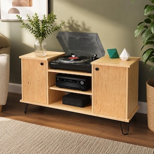 May include: A light wood media console with two side cabinets, a central open shelf, and black metal hairpin legs. A black turntable with a clear lid sits on top, alongside a glass vase with green foliage and two geometric decorative objects.