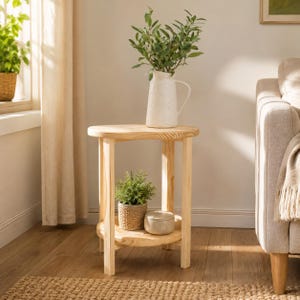 May include: A light-colored wooden side table with a round top and a lower shelf. The top holds a white textured pitcher with green leafy branches. The lower shelf holds a small potted green plant in a woven basket and a round beige container.