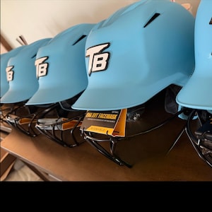 May include: A row of light blue batting helmets with black metal face masks. Each helmet features a white and black TB logo on the front and a yellow and black tag that reads EVO XVT FACEMASK and EVOSHIELD attached to the face mask wire.