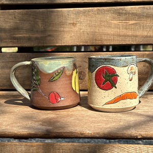 May include: Two ceramic mugs with handles. The left mug is brown with a green rim and features painted vegetables including a radish, pea pod, and yellow squash. The right mug is cream with a blue rim and features a red tomato, mushroom, and carrot.