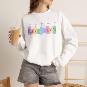 May include: A white long-sleeve crewneck sweatshirt featuring the text NAIL TECH in dark gray letters. Each letter is positioned above a colorful rectangular block topped with a small flag in shades of purple, yellow, teal, blue, and pink.