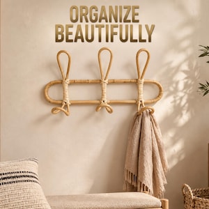 May include: A light brown rattan wall-mounted coat rack with three vertical loops and three lower hooks. The text "ORGANIZE BEAUTIFULLY" is displayed in gold lettering above the rack. A beige textured fabric hangs from the rightmost hook.