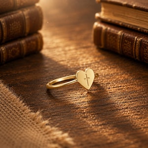 May include: A gold-toned ring featuring a heart-shaped centrepiece with a small, engraved cross symbol centred on the heart. The band is thin and smooth, reflecting light across its polished surface, creating a simple and clear design.