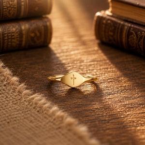 Könnte beinhalten: Ein goldfarbener Ring mit einer glatten, polierten Oberfläche und einem kleinen, mittig eingravierten Kreuzsymbol auf der Vorderseite. Der Ring hat eine schmale Schiene, die in ein breiteres, abgerundetes Mittelstück übergeht.