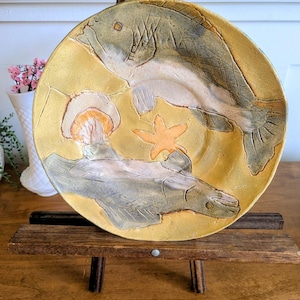 May include: A round ceramic plate with a textured yellow surface featuring two embossed fish figures in shades of grey and white, along with a small orange starfish. The plate is displayed on a wooden easel stand, showcasing detailed relief work.