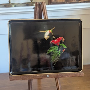 May include: A rectangular black serving tray with a gold-toned rim. The center of the tray features a detailed illustration of a hummingbird with green and orange plumage hovering over a vibrant red hibiscus flower with green leaves.