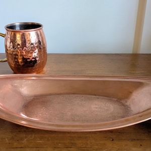 May include: A copper-colored hammered metal mug with a gold-toned handle and a matching elongated, shallow oval copper tray. Both items feature a metallic finish and are positioned together, showcasing their reflective surfaces and distinct shapes.