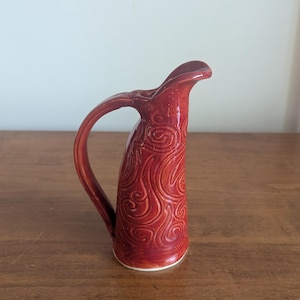 May include: A tall, slender ceramic pitcher with a glossy, deep red glaze. The exterior surface features a continuous, raised relief pattern of swirling, organic lines. It has a curved handle on one side and a prominent, elongated pouring spout.