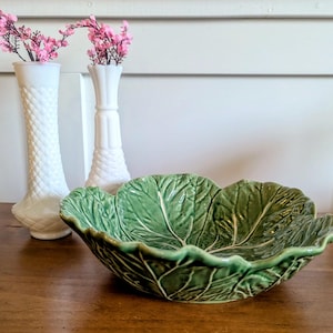 May include: A green ceramic bowl shaped like a cabbage leaf with prominent white veins and ruffled edges, accompanied by two white milk glass vases featuring a diamond-patterned texture, each holding small sprigs of pink artificial flowers.