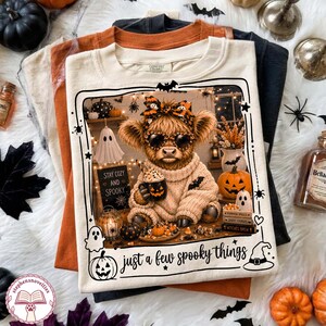 May include: A cream-coloured t-shirt featuring a graphic of a fluffy highland cow wearing leopard print sunglasses and a matching bow, holding a pumpkin mug. The design includes a ghost, a jack-o'-lantern, and the text "STAY COZY AND SPOOKY" and "just a few spooky things".