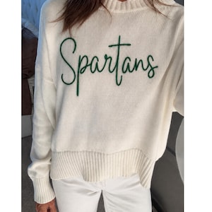 May include: A cream-colored long-sleeve knit sweater featuring the word Spartans embroidered in dark green cursive script across the chest. The sweater has a ribbed crew neckline, ribbed cuffs, and a ribbed hemline. The text Pictured - Size Small is visible.