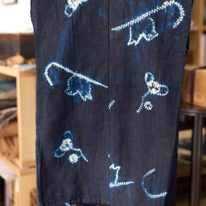 Peut inclure: Un textile bleu marine foncé orné de motifs floraux et de vignes blancs réalisés par la technique de teinture par réserve shibori. Le tissu présente une couture verticale centrale et un tissage texturé, avec des nuances d'indigo variées.