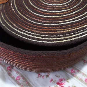 May include: A round, lidded storage basket constructed from coiled dark brown and metallic copper rope. The lid features a central decorative brown nut-like accent and a small leather-colored tag on the side. The basket has a textured surface.