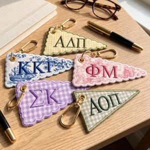 May include: Five triangular fabric keychains with gold-tone lobster clasps. The keychains feature embroidered Greek letters: AΔΠ on yellow gingham, KKT on blue toile, ΦM on pink toile, ΣK on purple gingham, and AOΠ on green gingham fabric.