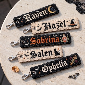 Op de afbeelding: Vijf rechthoekige geborduurde sleutelhangers met geschulpte randen en zilveren karabijnsluitingen. De sleutelhangers tonen de namen Raven, Hazel, Sabrina, Salen en Ophelia in gotisch lettertype, versierd met motieven zoals manen, vleermuizen, sterren, een heksenhoed, een toverdrankflesje, een kat en een sleutel.