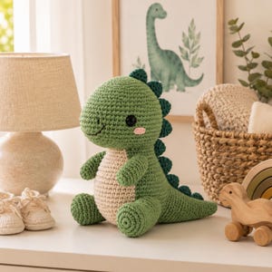 May include: A green crocheted dinosaur plush toy with a cream-coloured belly, dark green dorsal spikes, and black safety eyes. The stuffed animal features a rounded head, short arms, and a long tail, crafted with visible yarn stitches throughout.