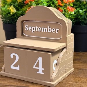 May include: A tan-colored perpetual block calendar featuring a rectangular sign with the white text September. Two square blocks display the number 24 in white font. The structure is made of a smooth material with a layered design and a flat base.