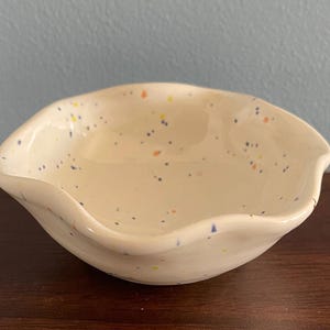 May include: A small, off-white ceramic bowl with a wavy, scalloped rim. The surface features a speckled pattern with tiny dots in shades of blue, orange, and yellow scattered across the interior and exterior of the glossy, rounded vessel.