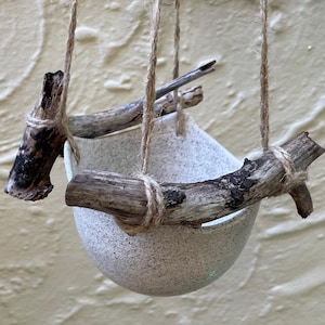 May include: A hanging planter featuring a speckled, off-white ceramic bowl. The bowl is suspended by twine and supported by two pieces of driftwood. The driftwood is tied to the bowl with twine. Suitable for indoor or outdoor use.