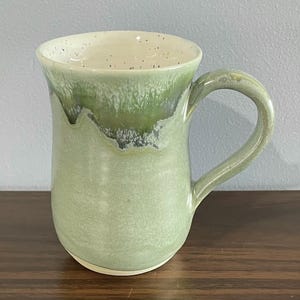 May include: A ceramic mug with a glossy finish, featuring a light sage green base that transitions into a darker forest green glaze near the rim. The interior is an off-white colour with small, dark speckles throughout the surface.