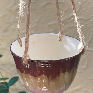 May include: Hanging ceramic planter with a cream interior and a gradient exterior of brown, tan, and pink. The planter is suspended by three jute ropes. A decorative piece for indoor or outdoor plants.