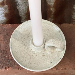 May include: A speckled off-white ceramic candle holder with a circular base, a central cylindrical socket, and a small side handle. A single plain white taper candle is inserted into the centre of the holder, which features a smooth glaze.
