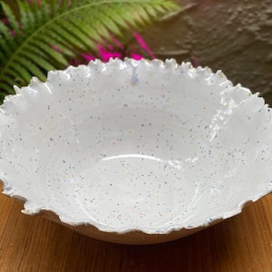May include: A white ceramic bowl with a speckled finish featuring small blue, orange, and yellow dots. The bowl has a distinctively uneven, ruffled, and textured rim that mimics the appearance of organic, jagged edges, contrasting with the smooth interior.