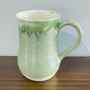 May include: A ceramic mug featuring a light green glaze that transitions into a darker, mottled green near the rim. The interior is off-white with dark speckles, and the mug has a curved handle and a smooth, unglazed white base.