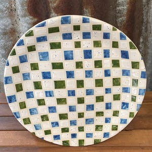 May include: A round ceramic plate featuring a chequered design of white, blue, and green squares. The plate has a speckled texture and a slightly raised edge. The pattern is evenly spaced across the surface.