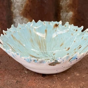 May include: A ceramic bowl with a light blue interior featuring brown streaks and a white exterior. The rim of the bowl has a jagged, leaf-like edge with a textured, irregular shape. The bowl has a glossy finish and a shallow, rounded base.