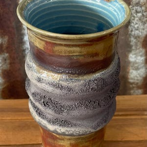 May include: Handmade ceramic tumbler with a textured, dark grey band around the middle. The upper portion features a gradient of brown, red, and gold, while the interior is a vibrant blue. The base is white.