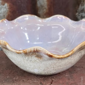 May include: A ceramic bowl with a ruffled rim. The bowl has a pearlescent interior and a textured, off-white exterior. The rim is edged with a band of brown. The bowl is sitting on a brick surface.