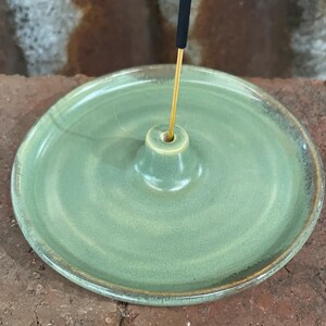 May include: A round, pale green ceramic incense holder with a central cone for the incense stick. The holder has a raised rim and a glossy finish. The incense stick is black and gold.