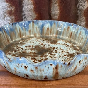 May include: A round ceramic pie dish with a scalloped edge, featuring a glossy glaze in shades of light blue, cream, and brown. The interior and exterior surfaces display a mottled, drip-style pattern with dark brown speckles and streaks.