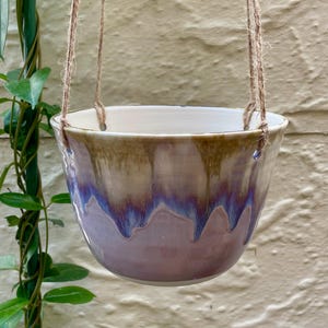May include: A ceramic hanging planter with a rounded bowl shape, featuring a glaze that transitions from a light cream at the top to a vibrant purple and soft pink at the base, suspended by three strands of natural brown jute twine.