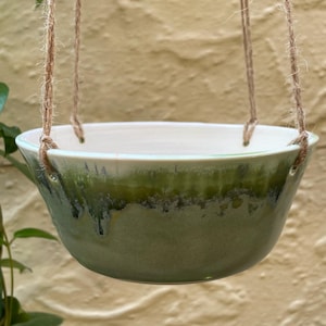 May include: A ceramic hanging planter bowl with a smooth white interior and a reactive green glaze on the exterior that drips down the sides. The bowl features three holes along the rim, threaded with natural jute twine for suspension purposes.
