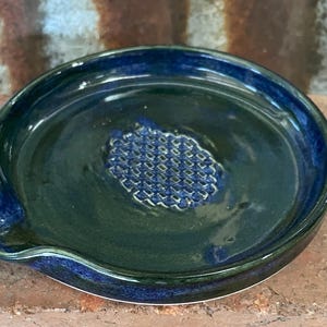 May include: A circular ceramic grater plate with a deep blue and black glossy glaze. The centre features a raised, textured diamond-patterned surface for grating ingredients, and the edge includes a small, integrated pouring spout.