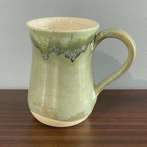 May include: A ceramic mug with a tapered body and a curved handle. The exterior features a light green glaze that transitions into a darker, mossy green drip pattern near the rim. The interior is a speckled off-white colour with a smooth finish.