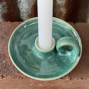 May include: A round, teal-coloured ceramic candle holder with a glossy, speckled finish and a small side handle. A single, plain white taper candle is inserted into the central socket of the holder, which features a wide, flat circular base.