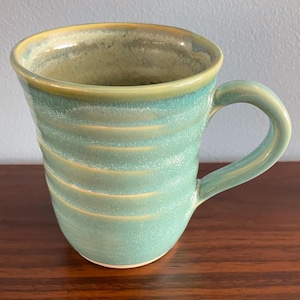 May include: A ceramic mug with a glossy finish in shades of teal and seafoam green. The exterior features horizontal ridges along the body, a curved handle on the right side, and a smooth, rounded rim with subtle yellow-toned glaze accents.