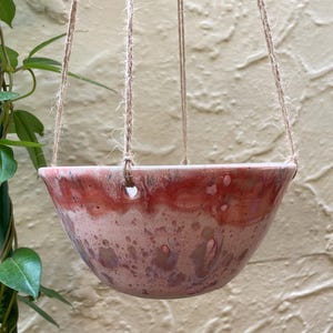 May include: A ceramic hanging planter bowl with a glossy glaze featuring a gradient of deep red at the rim transitioning into a mottled pink and light purple base. The bowl is suspended by three strands of natural jute twine through holes.