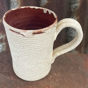 May include: A ceramic mug with a speckled off-white exterior featuring a horizontal ribbed texture. The interior of the mug is a deep reddish-brown colour, with the glaze dripping slightly over the top rim onto the outer surface of the vessel.