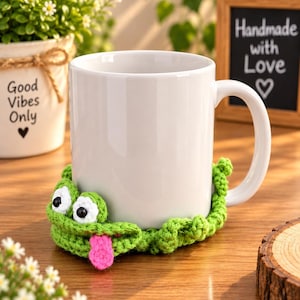 May include: A plain white ceramic mug featuring a green crocheted frog cozy wrapped around its base. The frog has large white and black eyes and a bright pink tongue sticking out from its mouth, adding a playful texture to the smooth mug.