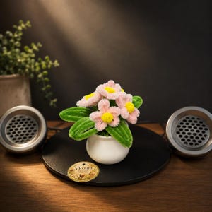 Pode incluir: Um pequeno vaso de cerâmica branca contendo um buquê de flores artesanais feitas de limpadores de cachimbo, com pétalas rosa claro e centros amarelo brilhante, acompanhadas por folhas verdes. Uma etiqueta redonda bege Vleoura Threads está anexada.