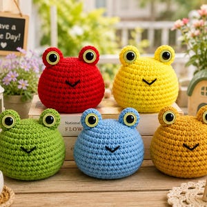 May include: Five round, crocheted frog plushies in red, yellow, green, blue, and orange. Each frog features two large, black-and-white plastic eyes on top and a simple black stitched V-shaped mouth. The toys are arranged on a wooden surface.