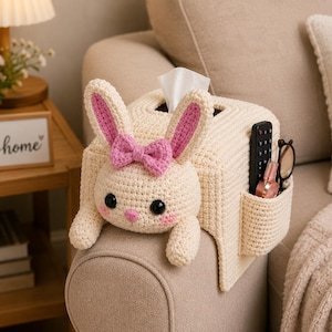 May include: A cream-colored crocheted tissue box cover shaped like a rabbit with pink inner ears, a pink bow, and black eyes. The cover features a side pocket holding a black remote control, a small pink bottle, and a pair of brown-rimmed glasses.