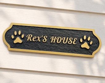 May include: A rectangular wall plaque with a textured black finish and gold-colored trim. The plaque features the text "Rex's HOUSE" in gold lettering, flanked by a gold paw print icon on both the left and right sides of the sign.